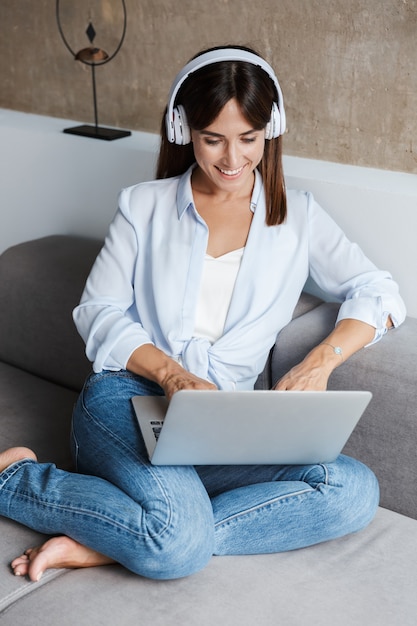 young positive happy woman indoors at home sit on sofa in living room listening music with headphones using laptop computer.