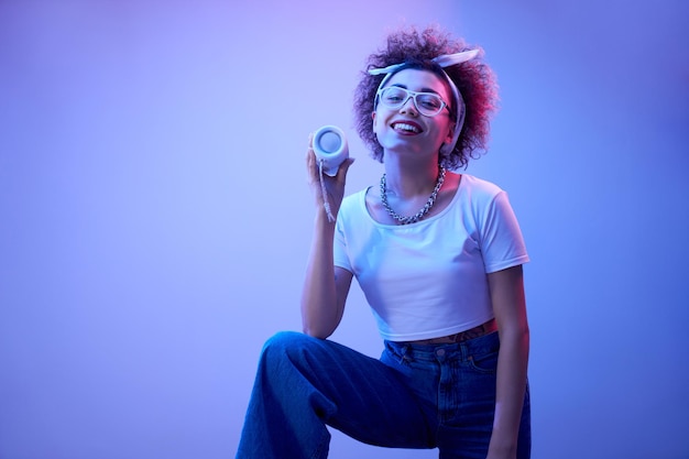 Young positive kazakh girl with afro hairstyle listens music in portable speaker dances and poses hip hop style