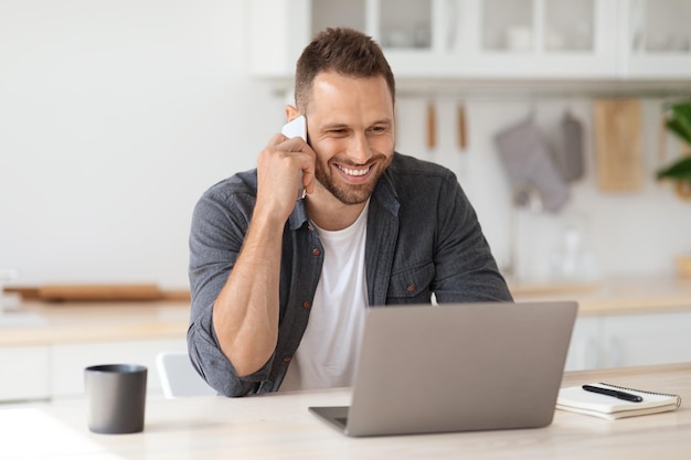 Young positive man freelancer working remotely at home looking at laptop and consulting client via cellphone