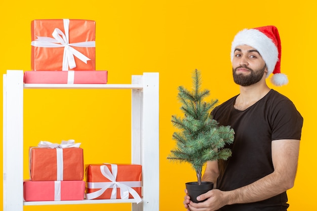 Young positive man with a beard and Santa Claus hat is standing near booth with gifts and Christmas tree on yellow wall. Concept of congratulation gift and discounts for Christmas