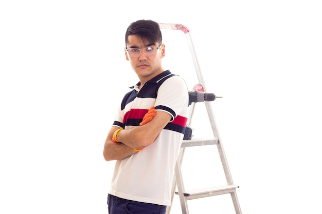 Young positive man with orange gloves standing near the ledder and holding electric screwdriver