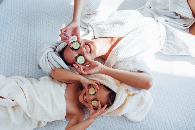 Young positive mother and daughter with towel on head lying on the bed together with cucumber.