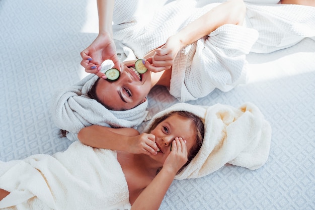 Young positive mother and daughter with towel on head lying on the bed together with cucumber.