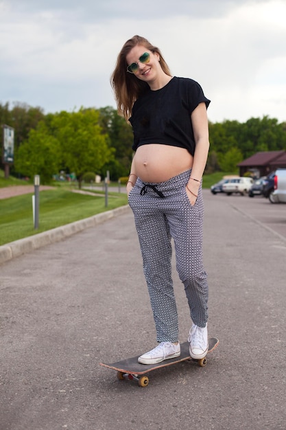 Young positive pregnant woman is skateboarding on the road on the background of green lawn