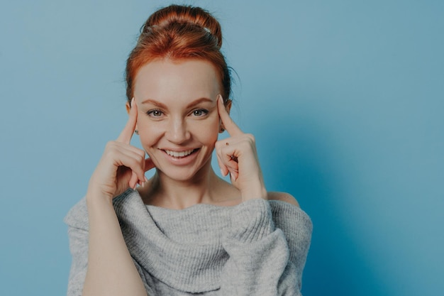 Young positive red haired woman with fingers on temples\
standing isolated on blue background