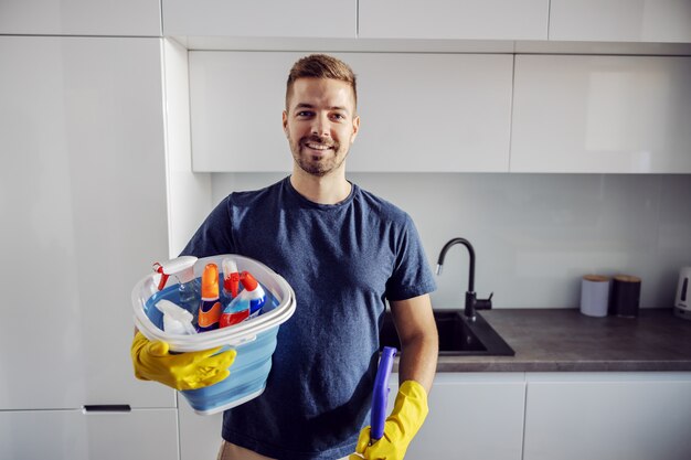 Giovane uomo barbuto degno sorridente positivo in piedi a casa con il secchio pieno di prodotti per la pulizia e si prepara a pulire tutta la casa.