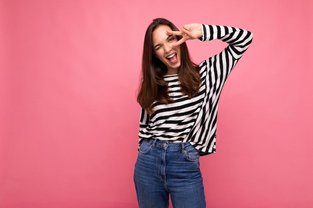 Young positive winsome happy beautiful brunette woman with sincere emotions wearing casual striped pullover isolated on pink background with copy space.