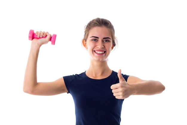 Young positive woman in blue T-shirt holding pink dumbbell and showing thumb up on white background in studio