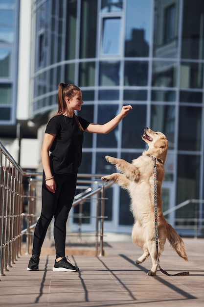 若いポジティブな女性は、ビジネスビルの近くで屋外を散歩するときに彼女の犬と一緒に楽しんでトリックをしています