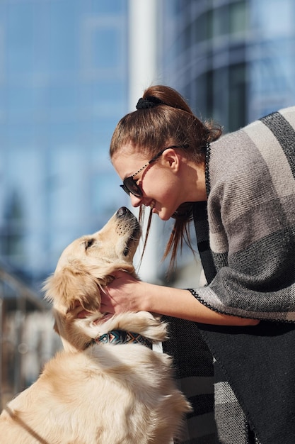 若いポジティブな女性は、ビジネスビルの近くで屋外を散歩するときに彼女の犬と一緒に楽しんでいます