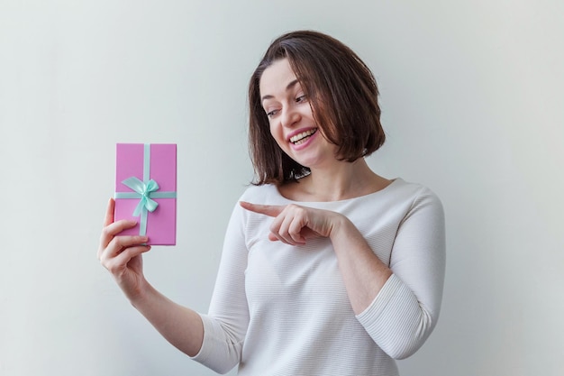 Молодая позитивная женщина, держащая небольшую розовую подарочную коробку на белом фоне, подготовка к празднику ...