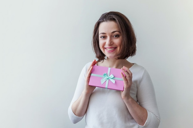 Young positive woman holding small pink gift box isolated on white background. Preparation for holiday. Girl looking happy and excited. Christmas birthday valentine celebration present concept