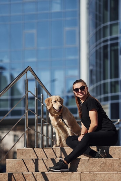 Young positive woman sitting on stairs with her dog when have a walk outdoors near business building
