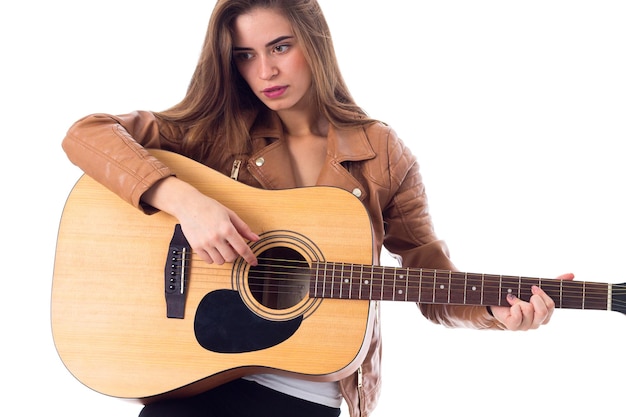 Young positive woman with long hair in brown jacket and black trousers holding a guitar in studio