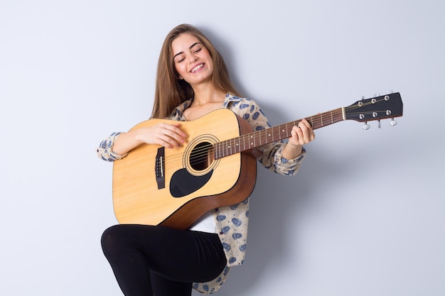 Young positive woman with long hair in brown jacket playing the guitar in studio