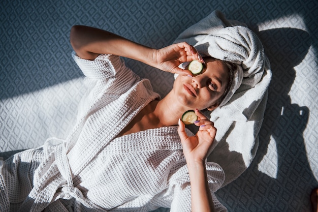 Young positive woman with towel on head lying on the bed with cucumber.