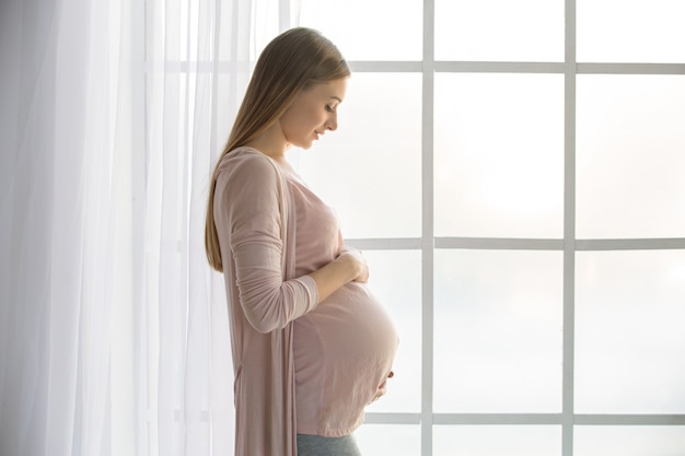 Giovane donna incinta che aspetta un genitore felice del bambino vicino alla finestra