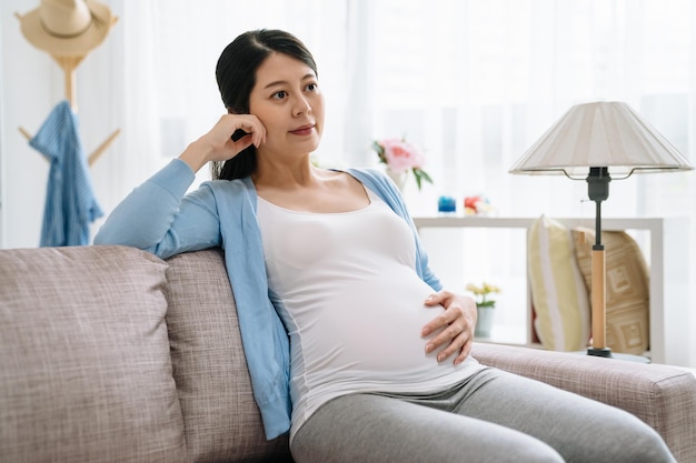 赤ちゃんの幸せな親を期待している若い妊娠中のアジアの女性。腹の子供の子供に触れる韓国の女性のお母さんの手。美しい女性は居心地の良いモダンな明るいアパートのソファソファのリビングルームで自宅でリラックスしました。