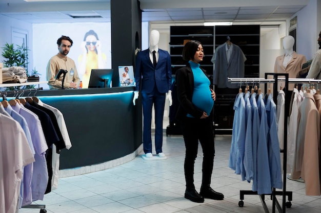 Young pregnant asian woman searching for maternity clothes in mall fashion boutique. Expectant mom standing near rack with hanging apparel in shopping center, choosing garment