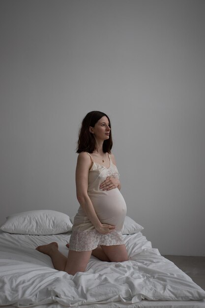 a young pregnant brunette girl in a silk nightgown sits on a bed