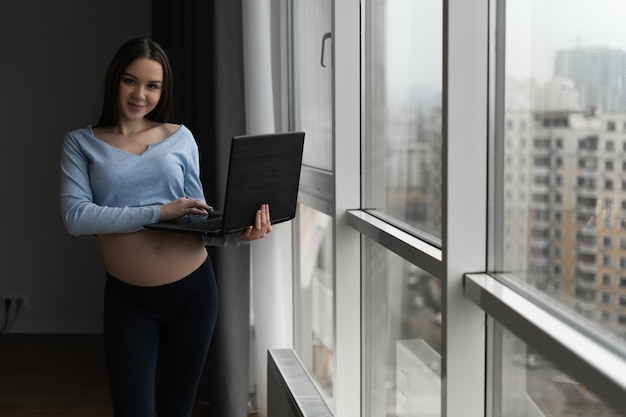 A young pregnant Caucasian woman stands near the window and works remotely behind her laptop