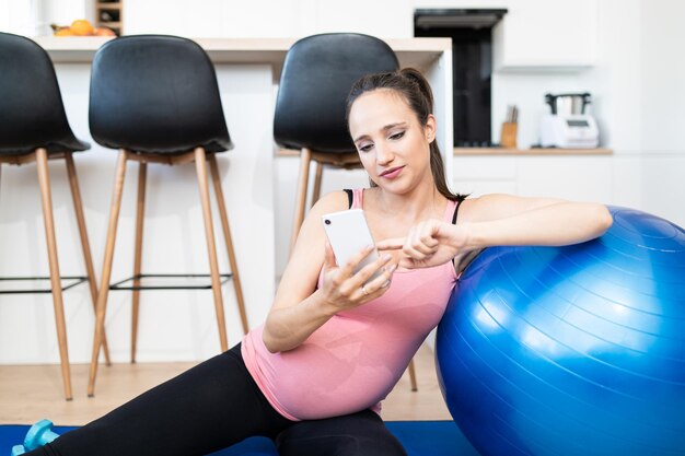 Young pregnant female using cell phone during working out in living room caucasian woman practicing