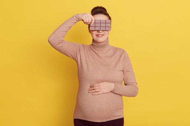 Young Pregnant female wearing casual clothing holding a bar of chocolate and covering eyes isolated over yellow wall.