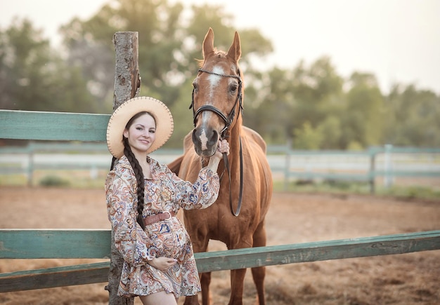 茶色の馬の隣の馬小屋で帽子をかぶった若い妊娠中の女の子