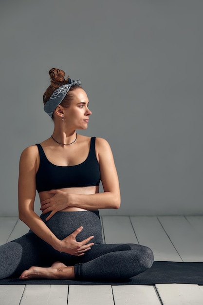 Young pregnant millennial woman sitting on the mat touches her\
belly after performing prenatal and meditation exercises at a yoga\
class concept of life and maternity