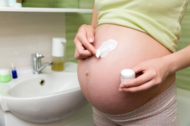 Photo young pregnant woman applying antistretch marks cream to the abdomen in the morning in the bathroom pregnancy maternity and bodycare