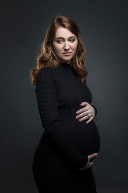 A young pregnant woman in a black tight dress touches her belly with her hands A beautiful pensive brownhaired woman is waiting for a baby Grey background Vertical