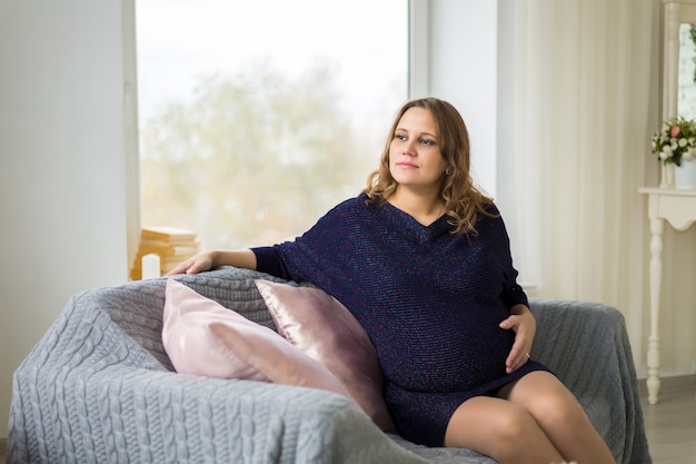 Photo a young pregnant woman in a blue dress is sitting by a large window with her hands on her stomach. waiting for a miracle