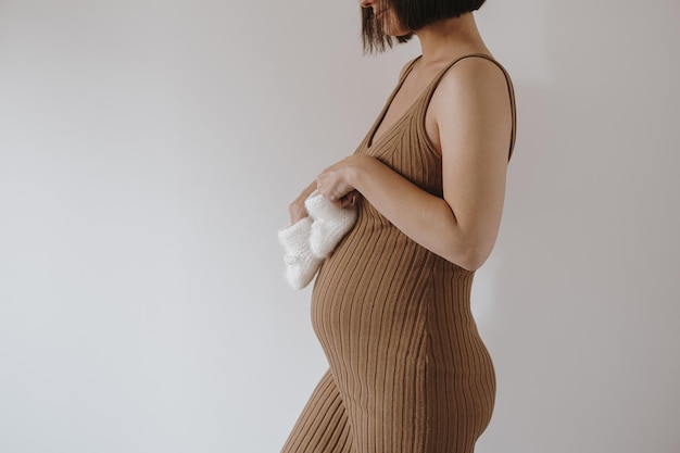 Young pregnant woman in brown dress hold white newborn socks against white wall Elegant aesthetic minimalist pregnancy maternity concept Baby expecting