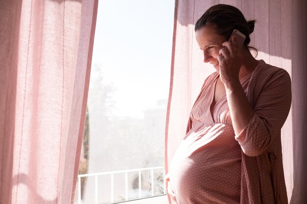 Young pregnant woman calling on the phone standing near window