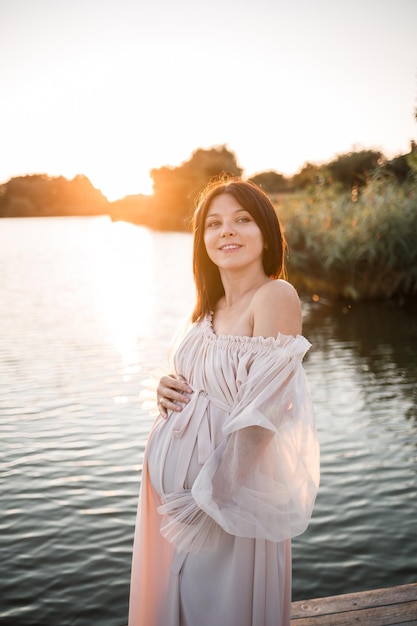 A young pregnant woman in a chiffon dress stands on a pier by the river against the backdrop of an orange sunset