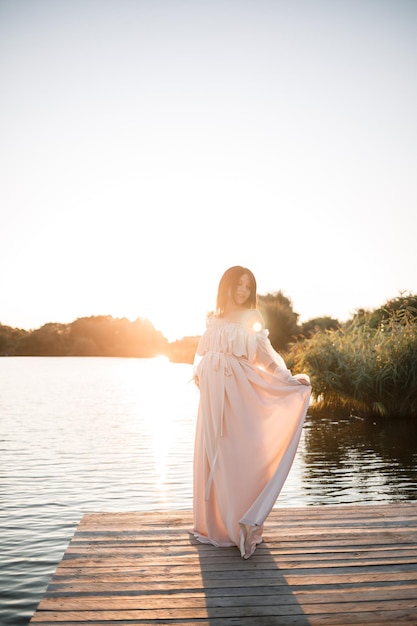 A young pregnant woman in a chiffon dress stands on a pier by the river against the backdrop of an orange sunset