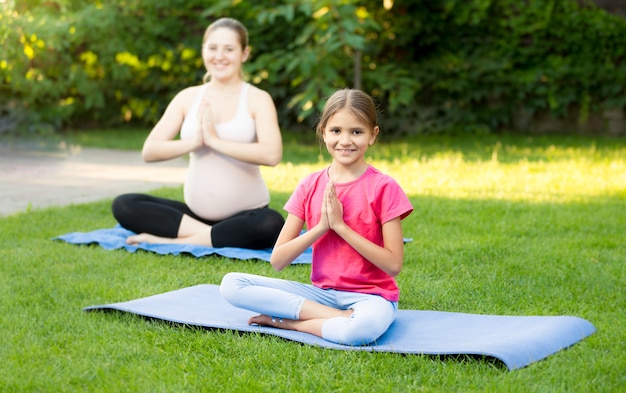 Young pregnant woman and cute girl practicing yoga on fitness mat at park