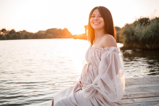 A young pregnant woman in a delicate dress by the river with a beautiful sunset Beauty health and gentle motherhood happy motherhood