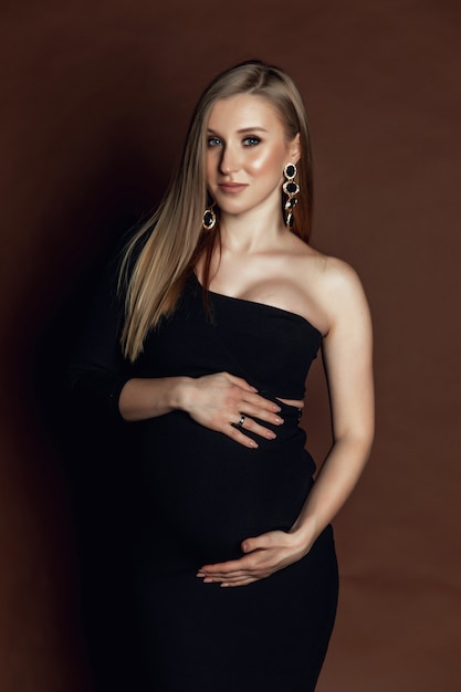 드레스에 젊은 임신 한 여자는 갈색 배경에 서있다. 사진 스튜디오에서 찍은 사진