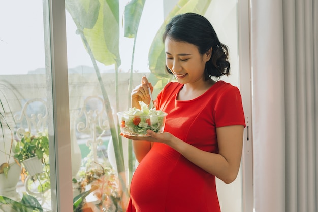 自宅の窓の近くで野菜サラダを食べる若い妊婦