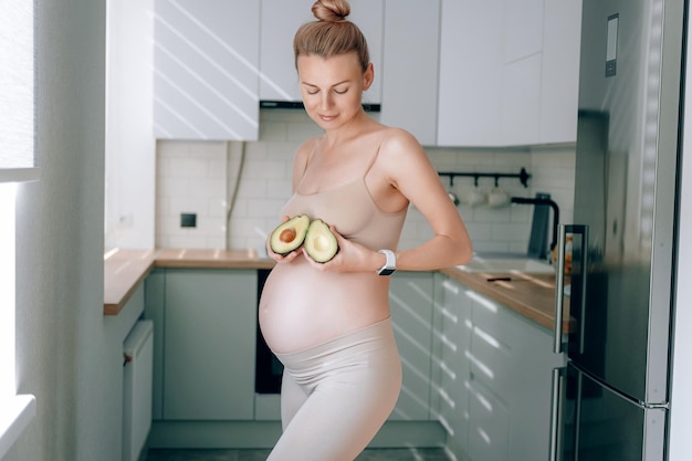 Photo a young pregnant woman of european appearance in her kitchen with an avocado in her hands the concept of proper nutrition of pregnant women for the correct development of the fetus