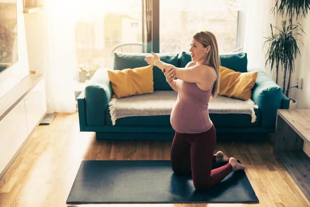 Young pregnant woman exercising at home. She workout on exercise mat and stretching in her living room.