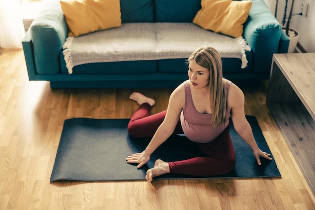 Young pregnant woman exercising mobility at home. She workout on exercise mat and stretching.