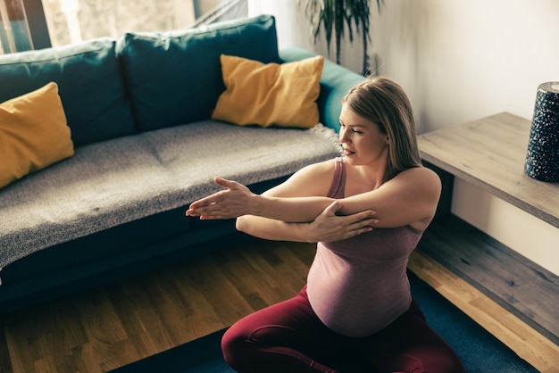 Young pregnant woman exercising yoga at home. She is doing stretching workout in her living room.