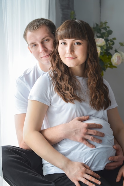 Photo young pregnant woman and her husband are hugging, touching the belly near window smiling