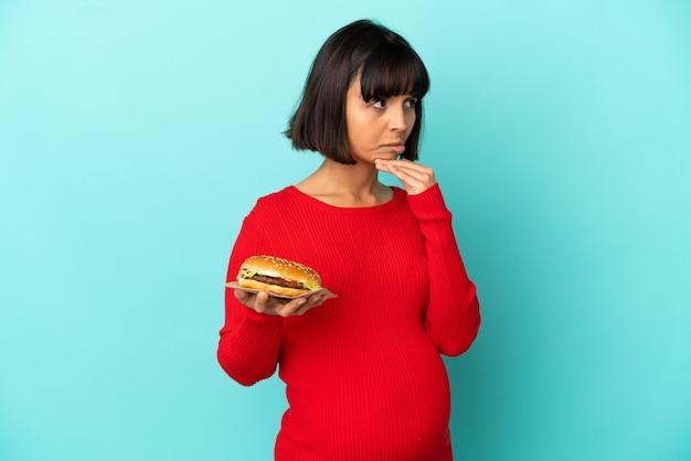 Young pregnant woman holding a burger over isolated background having doubts