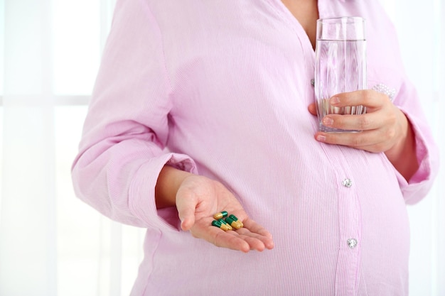 Young pregnant woman holding glass of water and tablets on light background