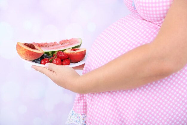 Young pregnant woman holding plate with fruits on bright surface