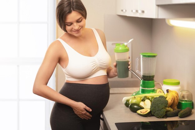 Giovane donna incinta che tiene in mano uno shaker con un cocktail proteico verde a casa