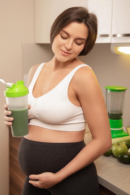 Foto giovane donna incinta che tiene in mano uno shaker con un cocktail proteico verde a casa